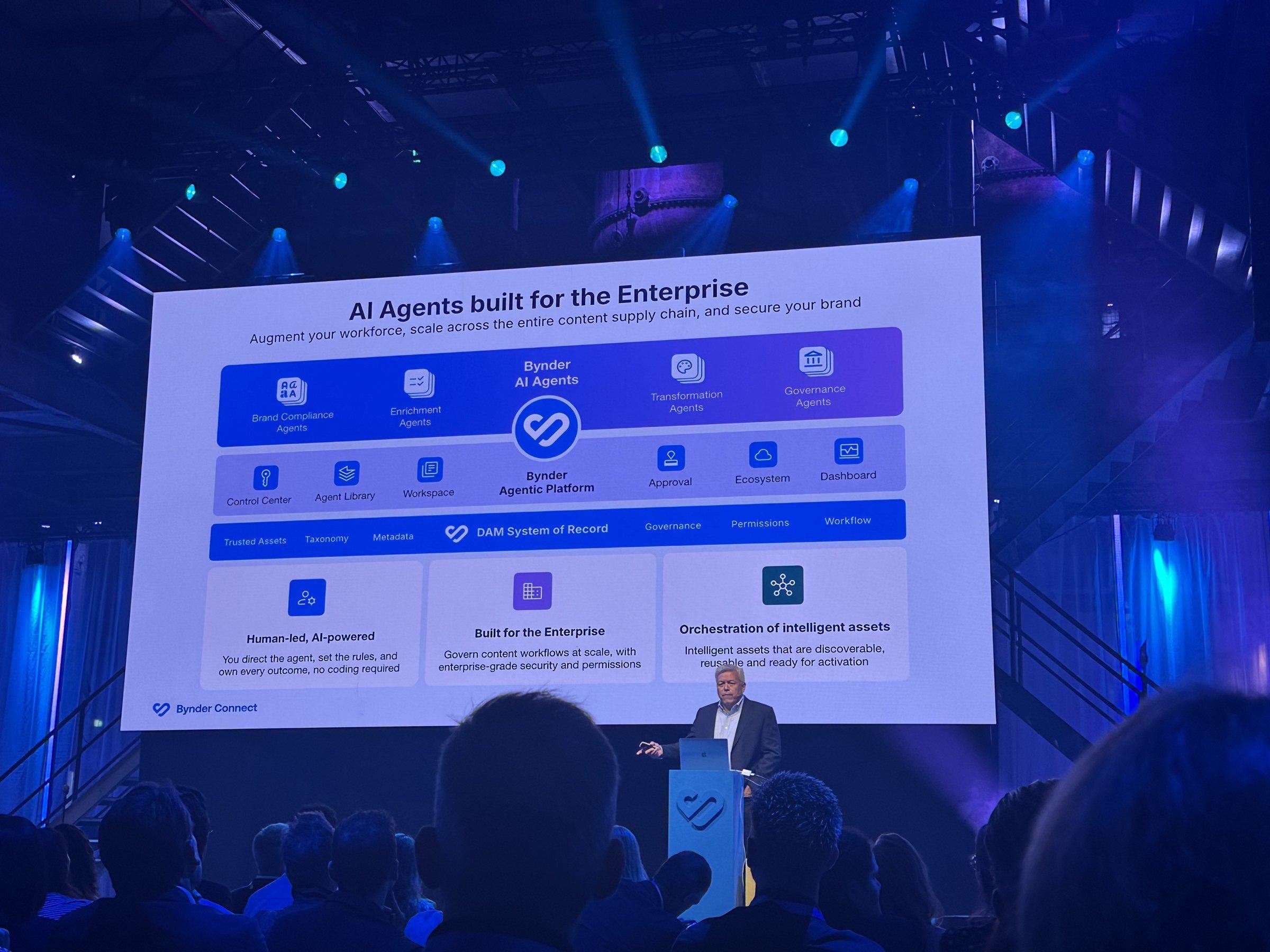 A Bynder speaker presents a slide titled AI Agents built for the Enterprise, showing brand compliance, enrichment, transformation, and governance agents.